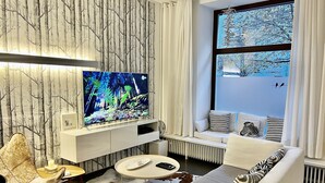 A modern living room with a large flat-screen TV, a sofa, a coffee table, and a window with a view of trees. - Best deal in town !  Cocoma Designer Apt deluxe in the center of Munich (Ludwigsvorstadt-Isarvorstadt)
