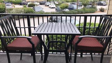 Parvekkeella on kaksi mustaa metallituolia ja pieni pöytä, ja sieltä avautuu näkymä parkkipaikalle, jossa on useita autoja, sekä taustalla olevaan rakennukseen.