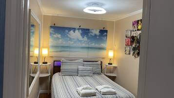 A bedroom with a bed, two bedside tables, a nightstand, a ceiling light, a wall-mounted lamp, and a large beach-themed painting.