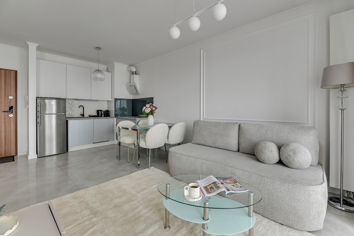 A modern living room with a grey sofa, a glass coffee table, and a dining area with white chairs.