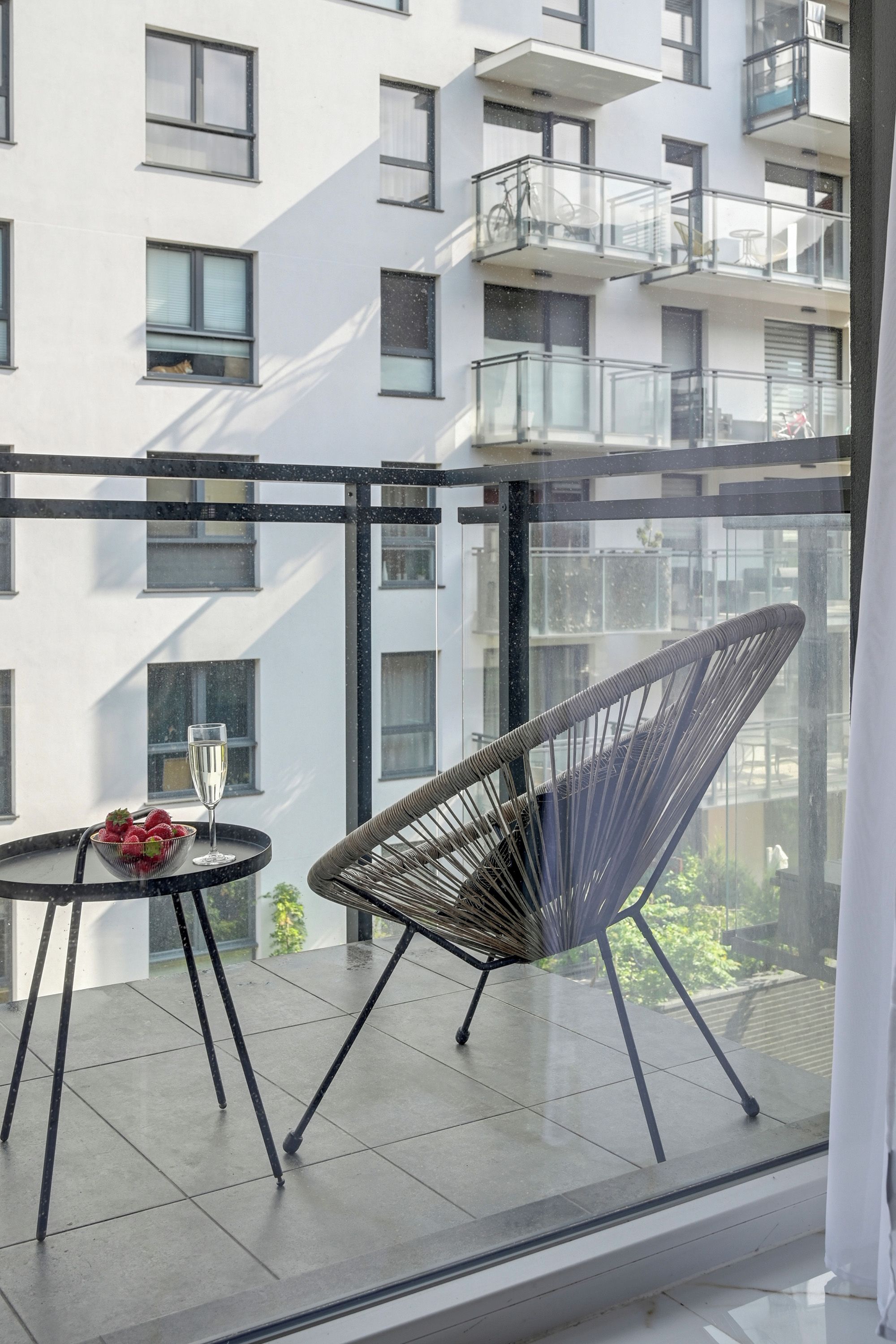 Un balcón con una silla, una mesa, baranda de vidrio y vista a un edificio con múltiples ventanas y balcones.