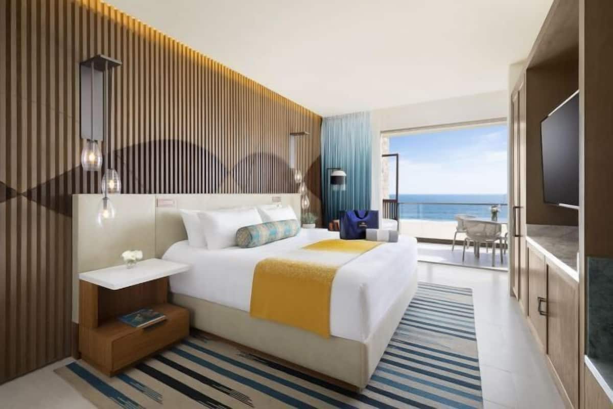 A modern hotel room with a bed, bedside table, lamp, and a view of the sea.