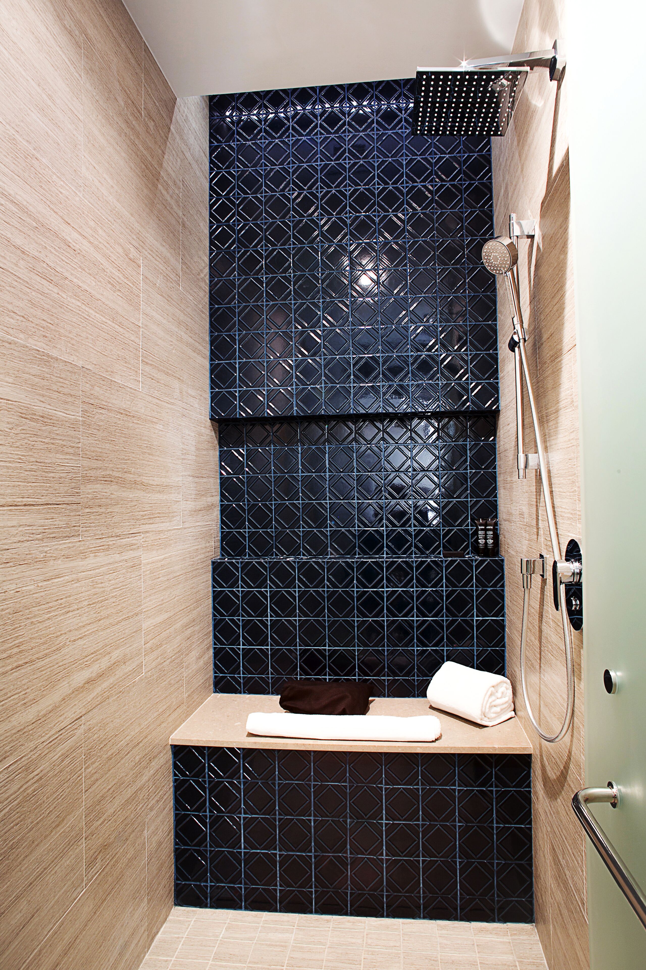 A modern shower with a black tiled wall, a wooden bench, and a handheld showerhead.