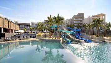 A swimming pool with a water slide, lounge chairs, palm trees, and hotel buildings in the background.