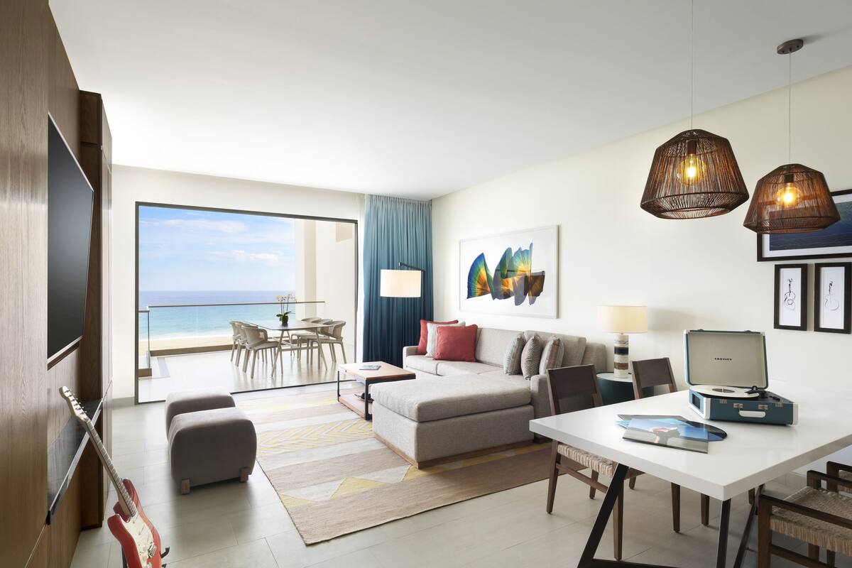 A modern hotel room with a large balcony, a sofa, a dining table, and a desk with a computer.