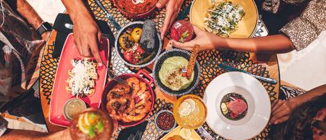 A group of people around a table with various dishes, including seafood, pasta, and a bowl of guacamole.