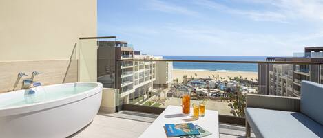 Un balcón con una bañera, una mesa con bebidas y vistas a la playa y edificios de hotel.