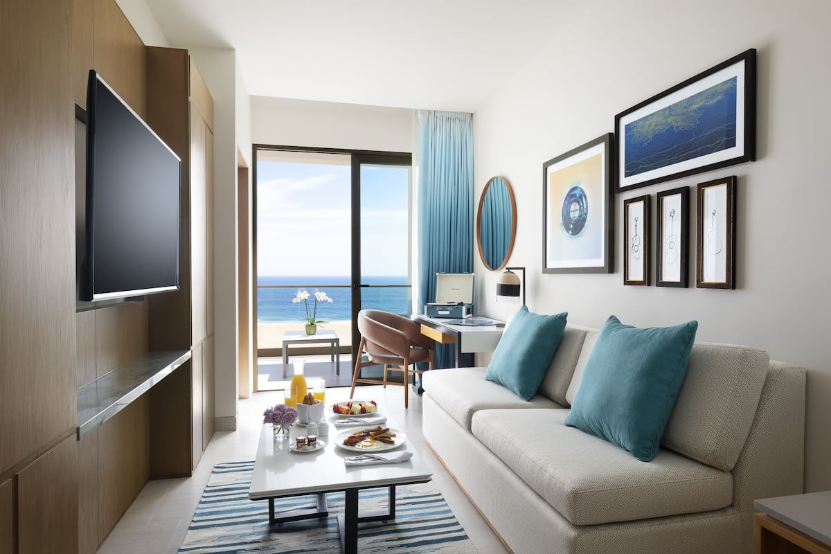 A modern hotel room with a large TV, a sofa with blue cushions, a dining table with breakfast items, and a view of the ocean through the sliding doors.