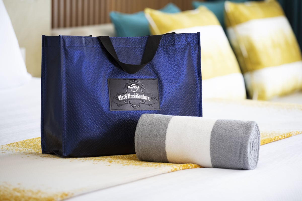 A blue tote bag with a label on a bed with white and yellow bedding.