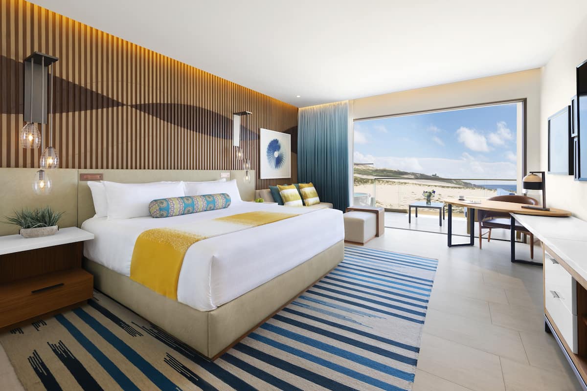 A modern hotel room with a large bed, a desk, and a view of the beach.