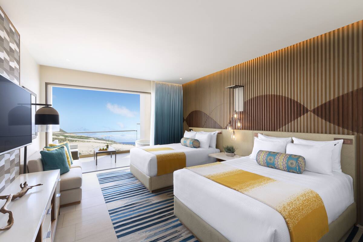 A hotel room with two beds, a TV, a desk, and a view of the beach.