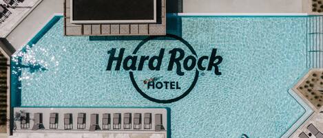 An aerial view of a hotel with a large swimming pool, lounge areas, and a central logo.