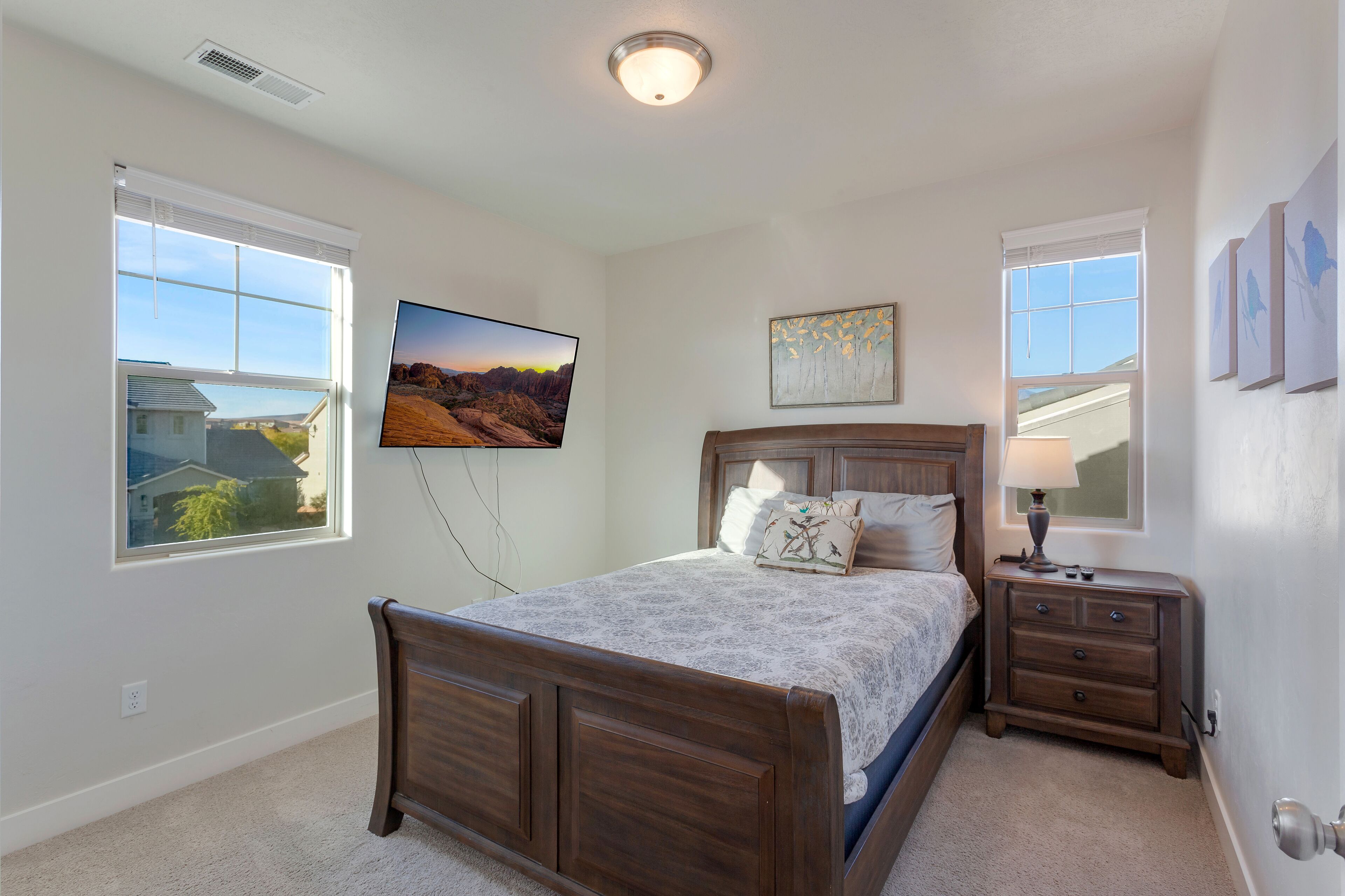 A bedroom with a wooden bed, a nightstand, a TV mounted on the wall, and two windows.