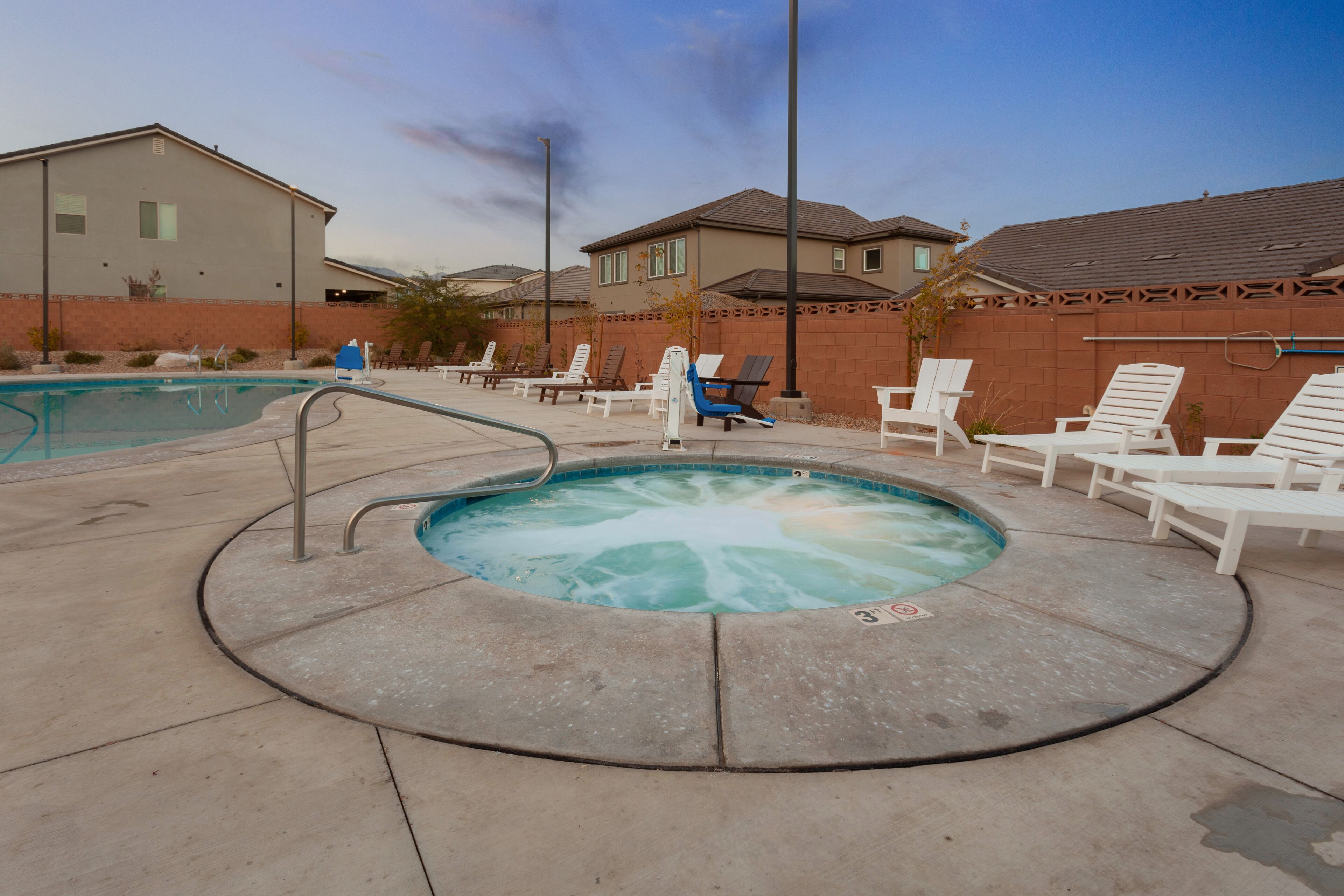 A hot tub surrounded by lounge chairs and a pool, with a brick wall and houses in the background.