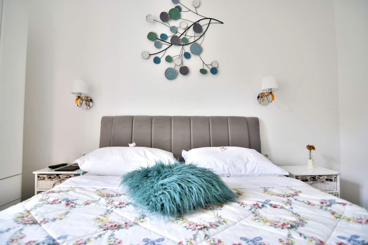 A neatly made bed with a floral-patterned bedspread, a grey upholstered headboard, and a decorative wall piece above it.