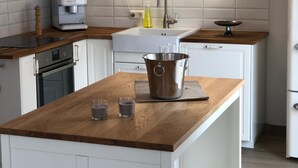 A modern kitchen with white cabinets, a wooden countertop, a built-in oven, a coffee machine, a sink, and a pendant light.