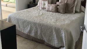 Un lit bien fait, avec une couvre-lit à motifs et plusieurs oreillers, une table de chevet avec une lampe, et une vue sur un balcon par une porte ouverte.