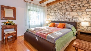 Une chambre avec un mur en pierre, des poutres apparentes, un lit avec une literie aux couleurs vives, une table de chevet avec une lampe et un banc en bois.