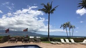 A pool area with lounge chairs, a flag, and a view of the ocean. -  'Island Casual' Oceanfront  'Elegantly Comfortable'  (Wailuku)