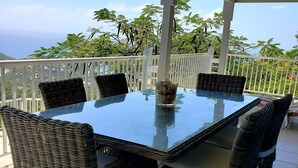 Un espace extérieur abrité, aménagé pour les repas, avec une table en verre et des chaises en osier, entouré d’une clôture blanche et d’une végétation luxuriante.