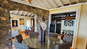 Une table ronde en verre, des chaises en osier, un mur en pierre et un meuble à vin dans une pièce dotée d’un plafond haut et de poutres apparentes.