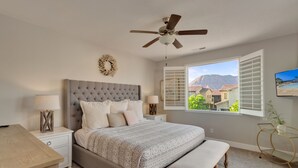 A bedroom with a tufted headboard, a large window with shutters, a ceiling fan, and a view of a mountain and buildings. - Paradise Village Resort 86, Private Hot Tub, Game System, Community Amenities, and more (Santa Clara)