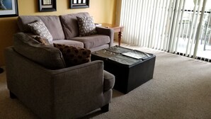 A living room with a sofa, armchair, and ottoman. There are framed pictures on the wall and a window with blinds. - One Bedroom, First Floor, Fully Furnished Condo (Kailua-Kona)