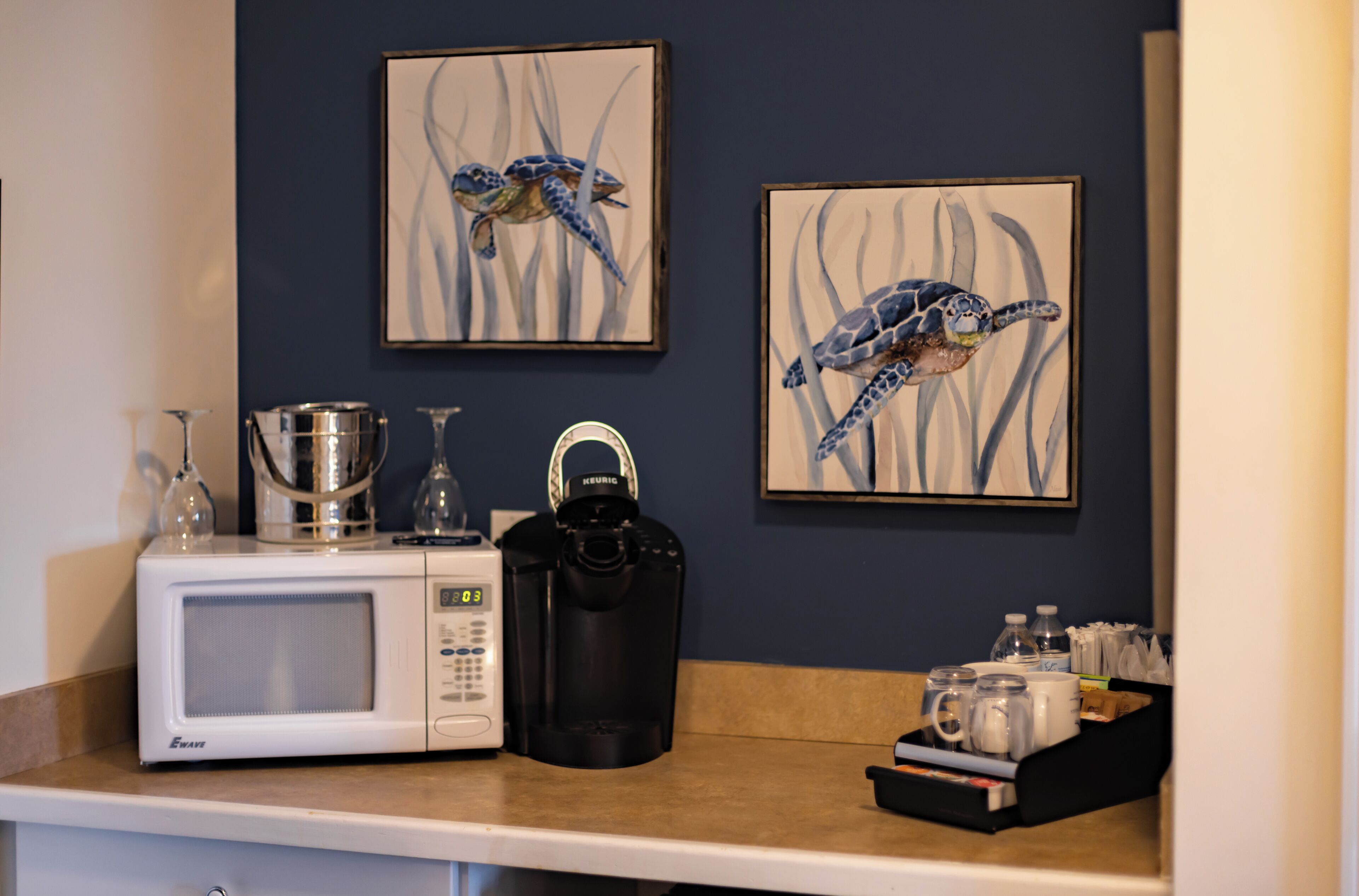 A kitchenette with a microwave, coffee maker, and a tray with cups and a kettle. There are two framed artworks of sea turtles on the wall.