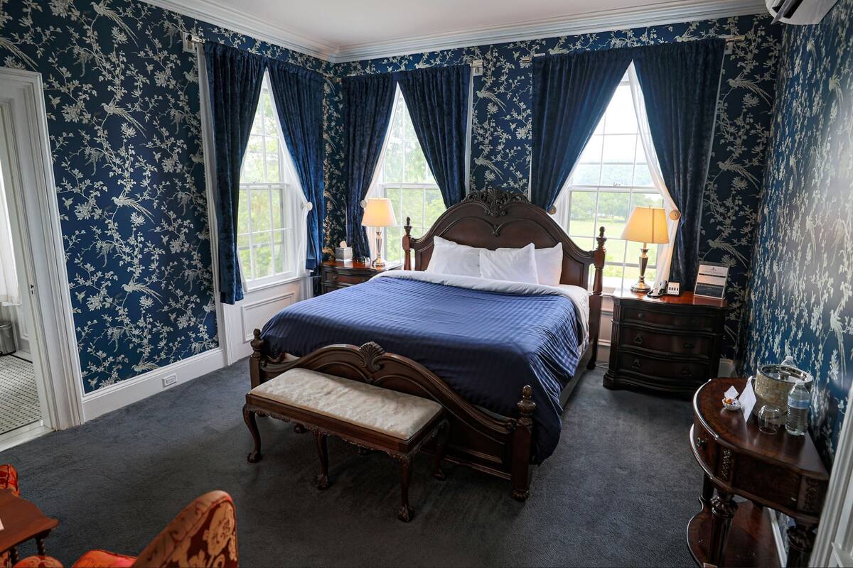 A bedroom with a large bed, a bench, a nightstand, a dresser, and two windows with curtains.