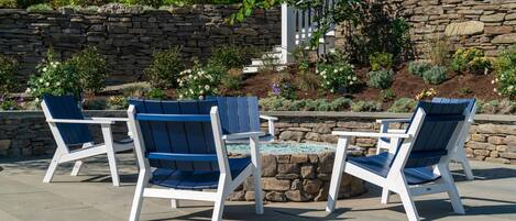 Un patio con cuatro sillas Adirondack y una mesita pequeña, rodeado por un muro de piedra y un jardín con diversas plantas y flores.