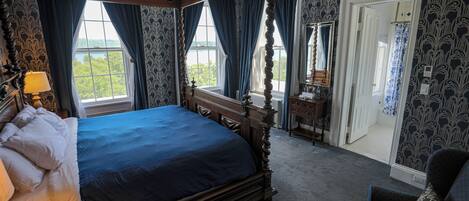 A four-poster bed with a dark blue bedspread, a wooden nightstand with a lamp, a dresser with a mirror, and a view of the outdoors through large windows.