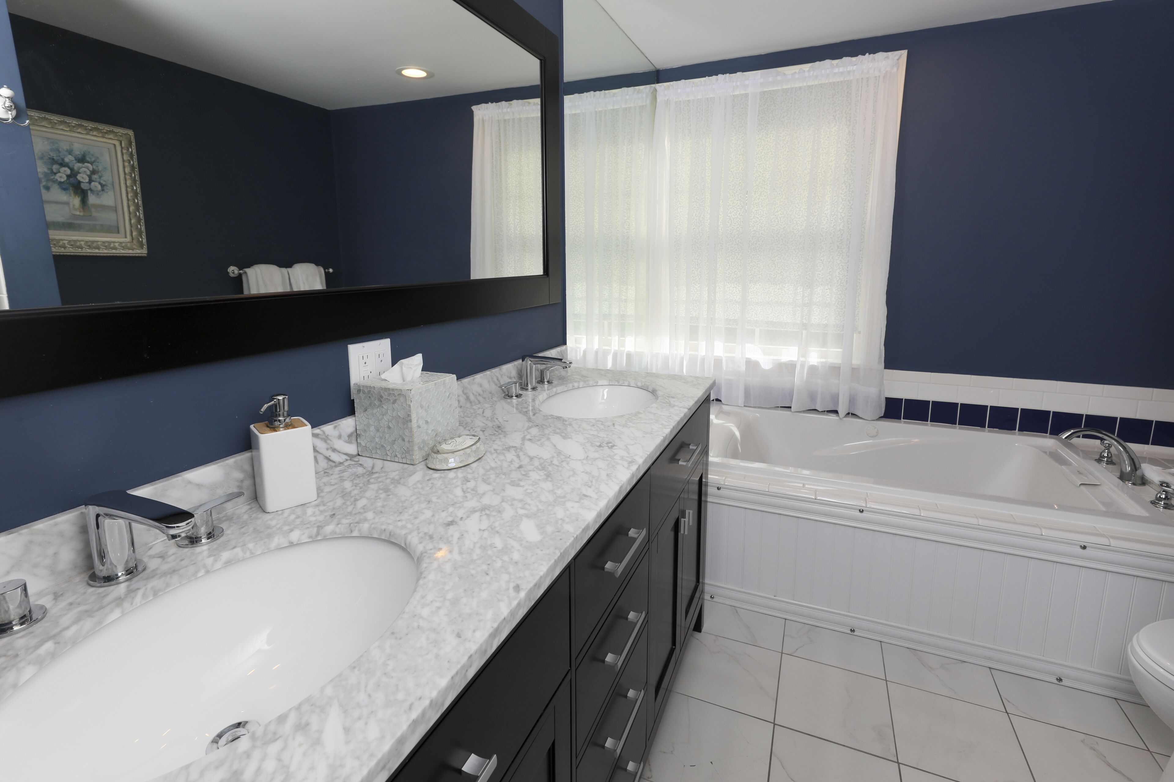 A modern bathroom with two sinks, a large mirror, a bathtub, and a window with sheer curtains.