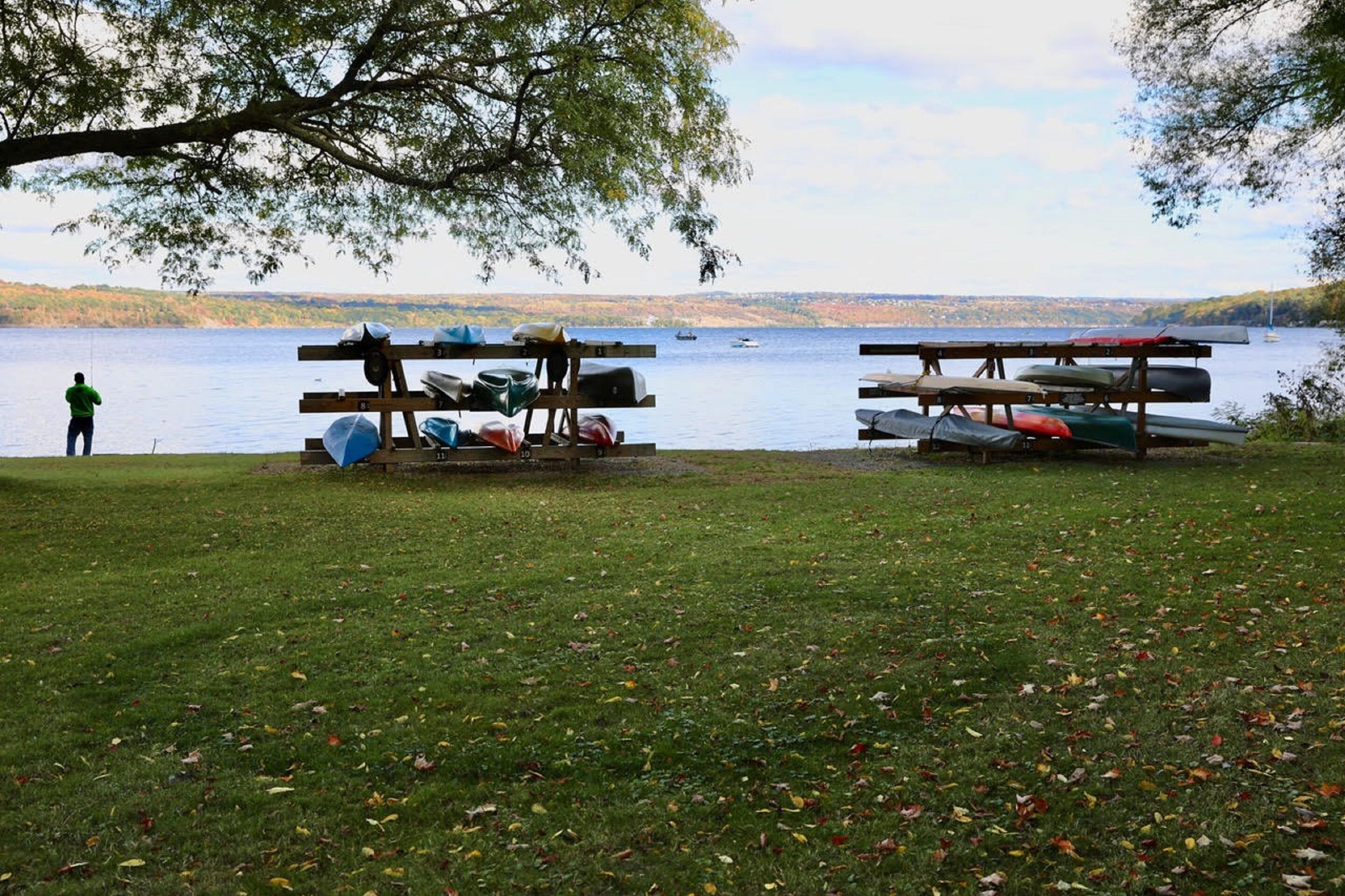 A lakeside setting with a dock and canoes, a person fishing, and a grassy area with scattered leaves.