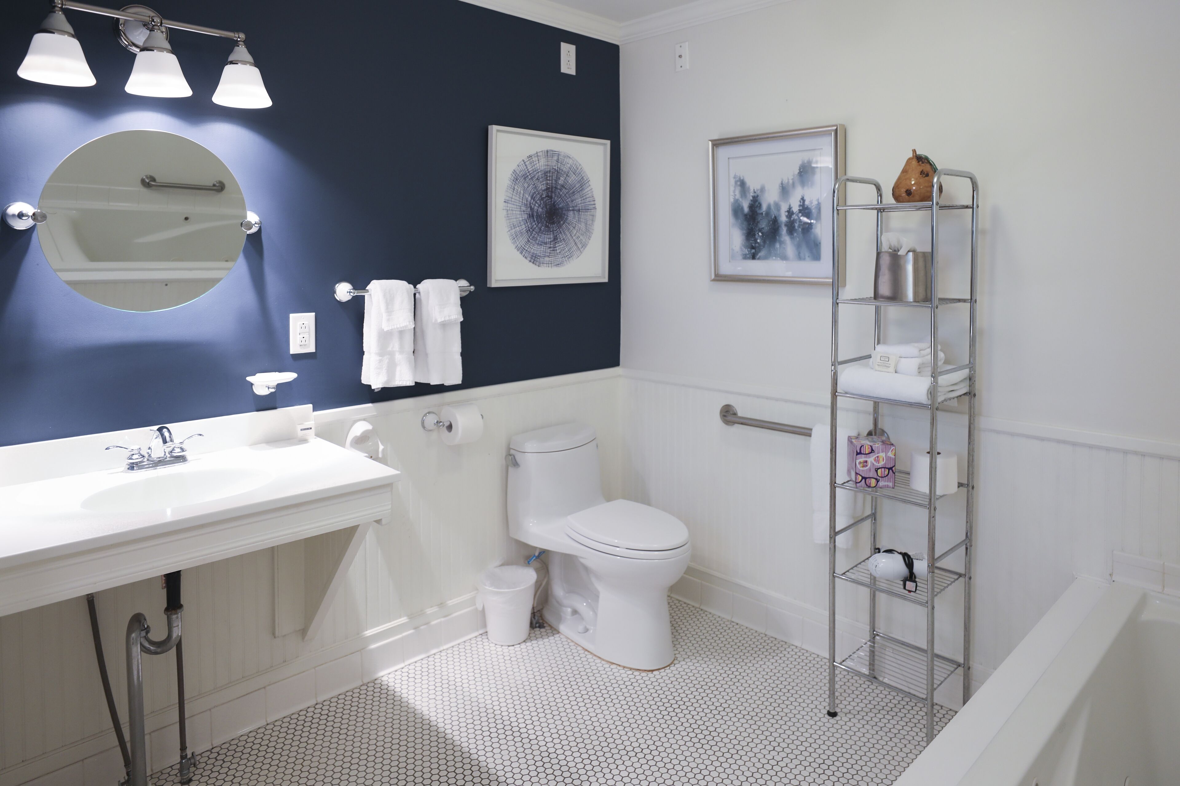 A bathroom with a white sink, round mirror, toilet, and a metal shelf with towels.