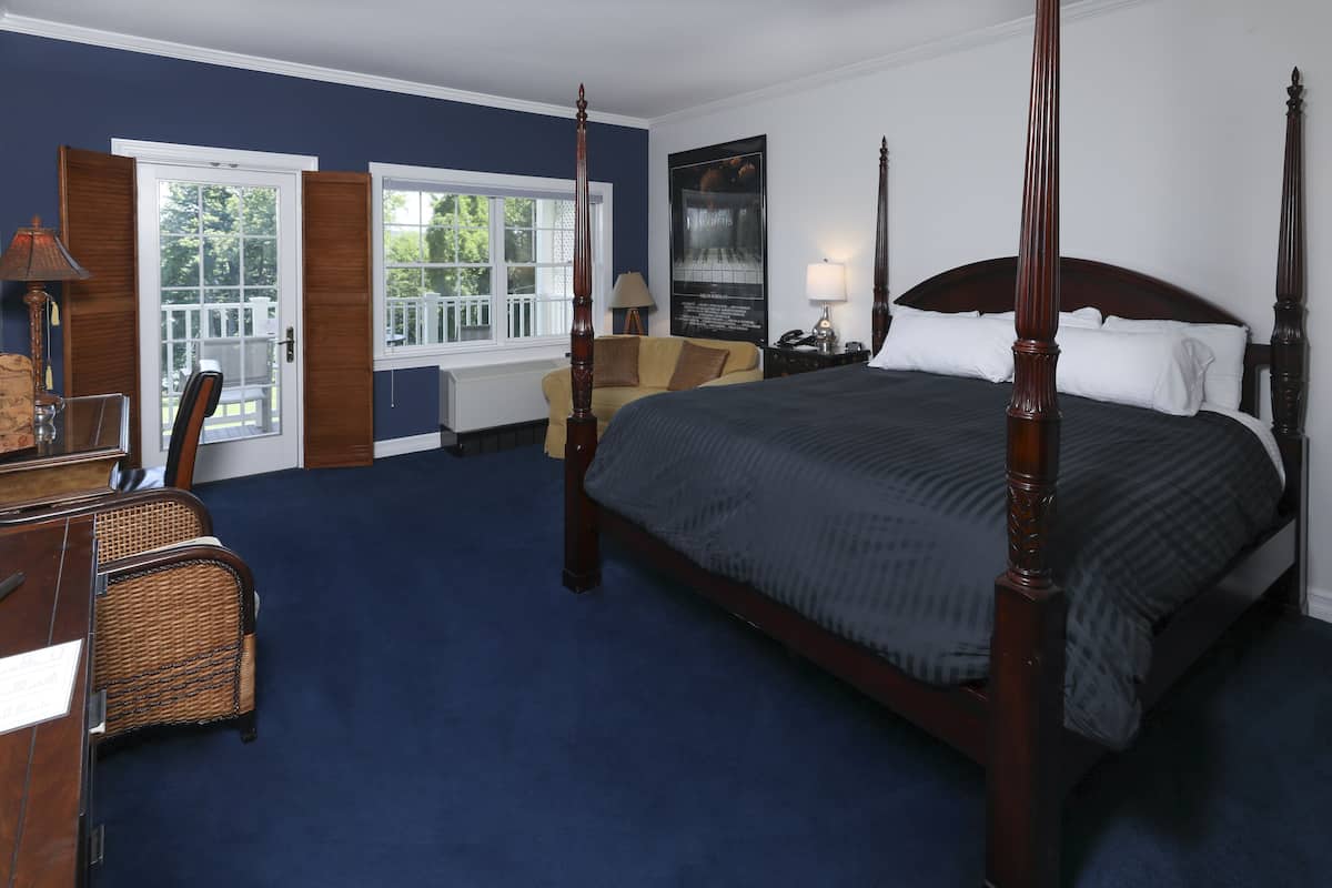 A four-poster bed with a dark headboard, a wooden desk with a chair, a small table with a lamp, and a window with a view of greenery.