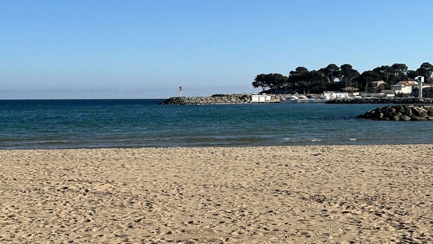 Una playa arenosa con un rompeolas rocoso, un faro y una hilera de edificios con árboles.
