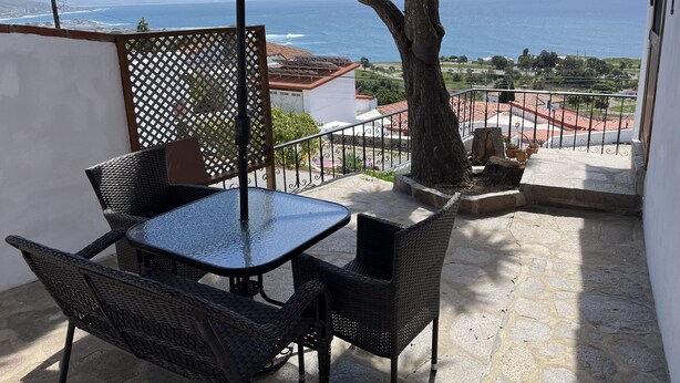 A balcony with a table and chairs, a red umbrella, and a view of the sea and distant mountains.