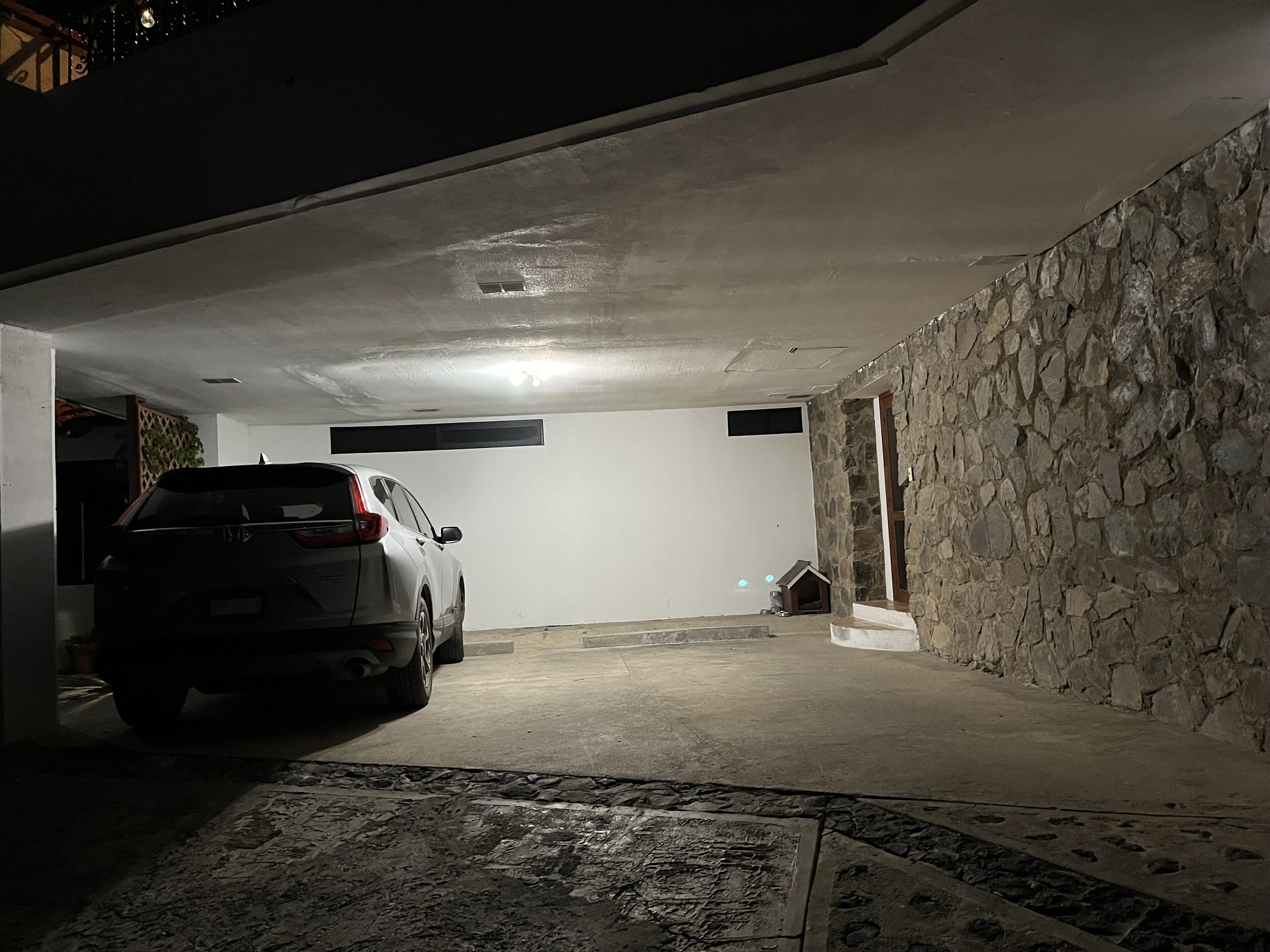 Un garaje tenuemente iluminado con un coche aparcado, un muro de piedra y una caseta pequeña para perro.