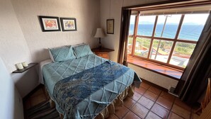 A bedroom with a bed, a nightstand, a lamp, and a window with a view of the sea.