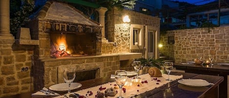 An outdoor dining area with a stone fireplace, a table set for two with wine glasses and plates, and a pergola overhead.