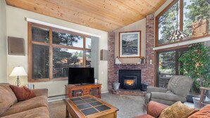 A cozy living room with a fireplace, large windows, and comfortable seating. - Woodlands 2 Bedroom & Loft Townhouse In A Beautiful Wooded Complex (Mammoth Lakes)