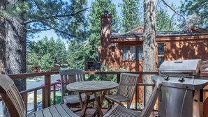 A wooden deck with a round table and chairs, a grill, and a view of a cabin surrounded by trees. - Woodlands 2 Bedroom & Loft Townhouse In A Beautiful Wooded Complex (Mammoth Lakes)