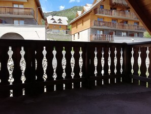 Ein hölzerner Balkon mit Steinbalustrade, der auf eine gebirgige Landschaft mit Gebäuden blickt.