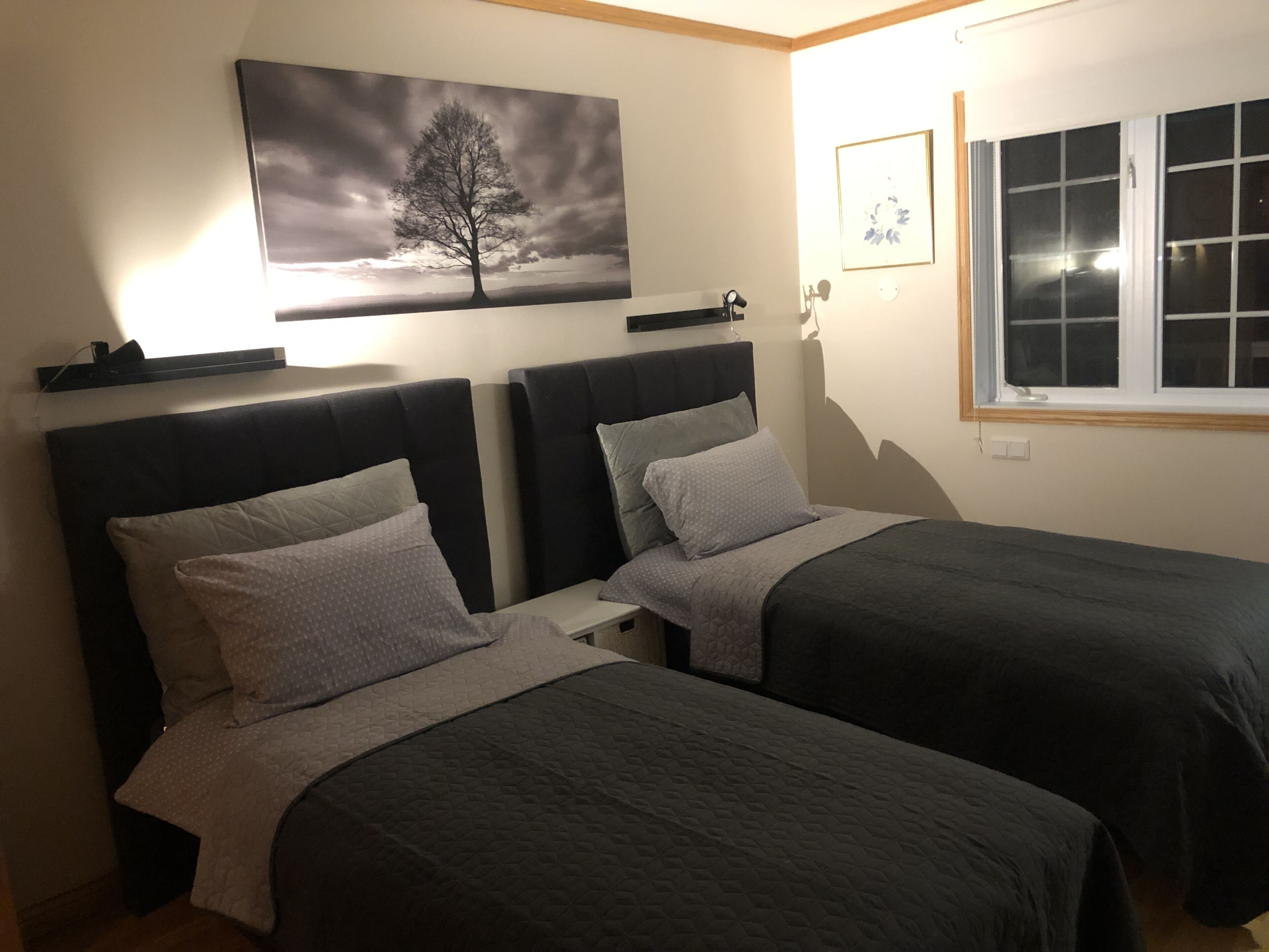 A bedroom with two beds, a painting of a tree, and a window with blinds.