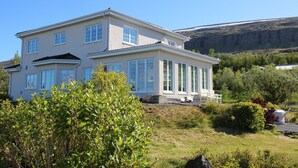 Une maison à deux étages, dotée de grandes fenêtres et d’un balcon, entourée de verdure et avec une chaîne de montagnes en toile de fond.