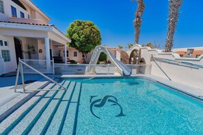 Une piscine en forme de dauphin, entourée d’une clôture blanche, de palmiers et d’un bâtiment doté d’un toit en tuiles.