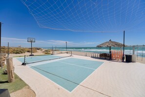A beach volleyball court with a net, a basketball hoop, and a thatched-roof structure.