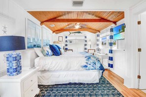 Un dormitorio con techo de madera, cama con ropa de cama azul y blanca, mesita de noche con lámpara y televisor en la pared.