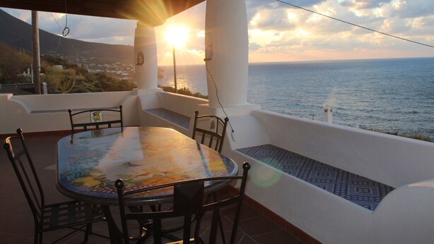 A dining area with a table and chairs, overlooking a coastal view with the sun setting over the ocean. - The casuzza of RE !! (Malfa)