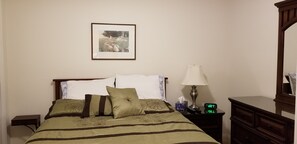 Ein ordentlich bezogenes Bett mit floralem Überwurf, eine Nachttischlampe, ein Nachttisch mit einer Uhr und ein gerahmtes Bild an der Wand.